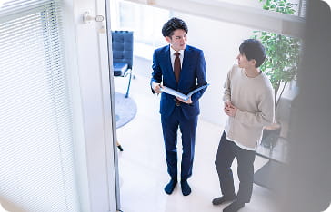 明るいオフィスの通路で、スーツ姿の男性が資料を手に説明している