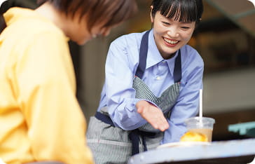 カフェのような空間で、エプロン姿の女性スタッフが笑顔で説明している